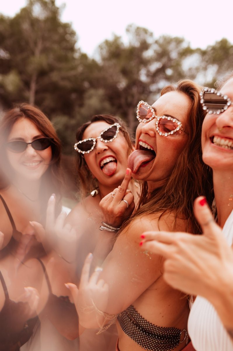 Vier Frauen lachen ausgelassen in die Kamera, Sonnenbrillen, Sommerlicht.